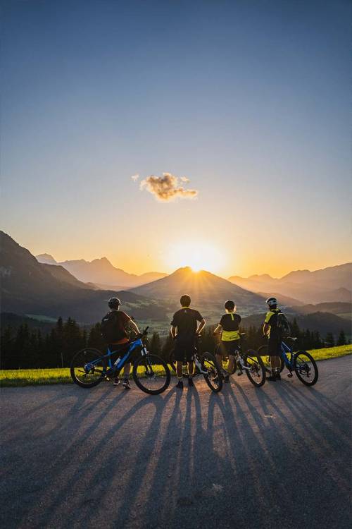 A group of cyclists stand on an asphalt road and admire the sunset in the Alps.