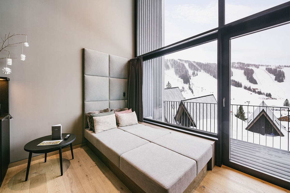 Sofa in the Superior Family Suite at the TRIFORÊT alpin.resort in front of a panoramic window overlooking the snowy Alps.