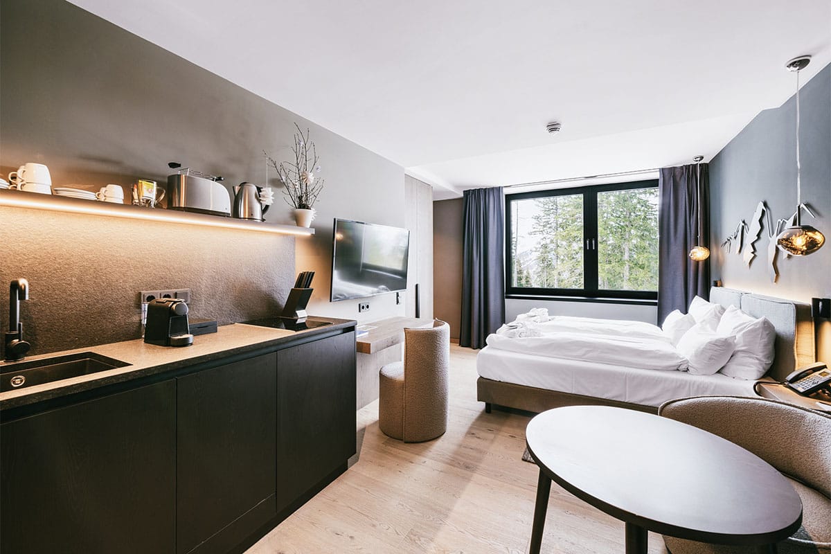 Bed, kitchenette and TV in the studio apartment at the TRIFORÊT alpin.resort with a view of the Hinterstoder landscape.
