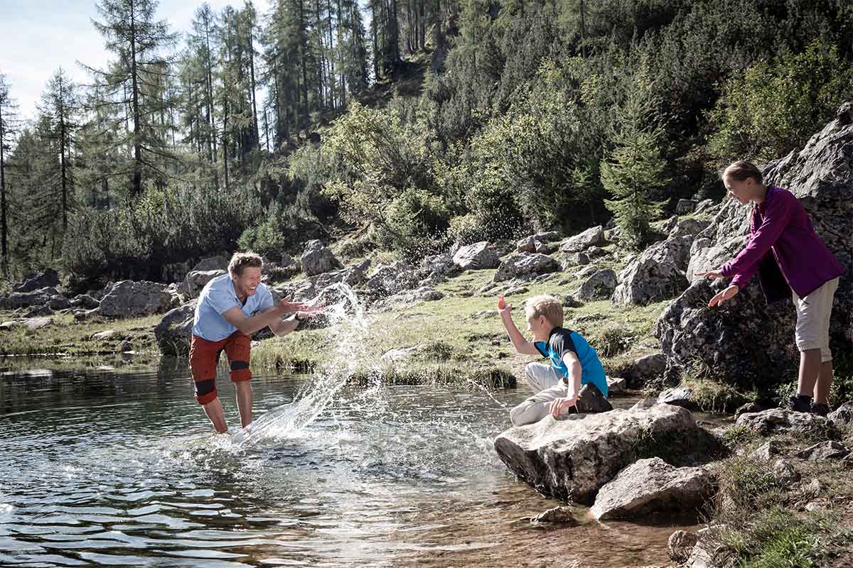 Familie spielt im glasklaren Wasser auf einer Wandertour durch die Alpen im Sommerurlaub im TRIFORÊT alpin.resort.