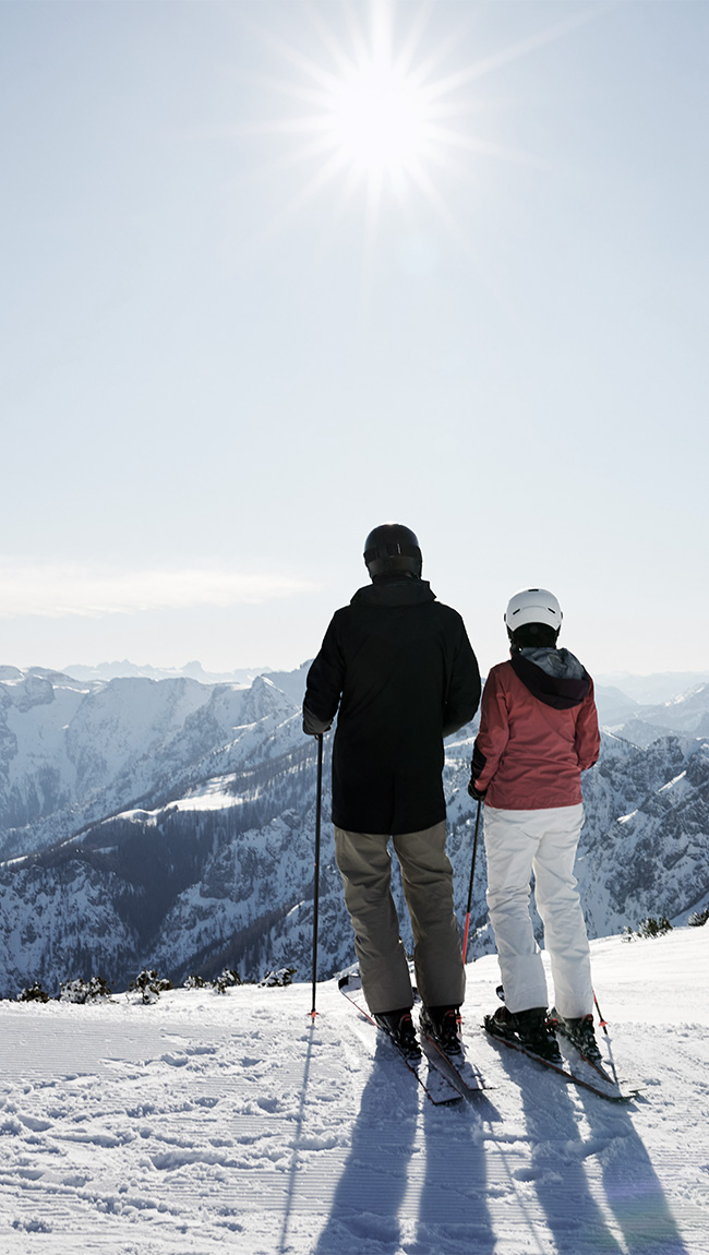 Ein Paar steht gemeinsam auf dem Berg in Skiern und blickt hinunter ins Tal von Hinterstoder.