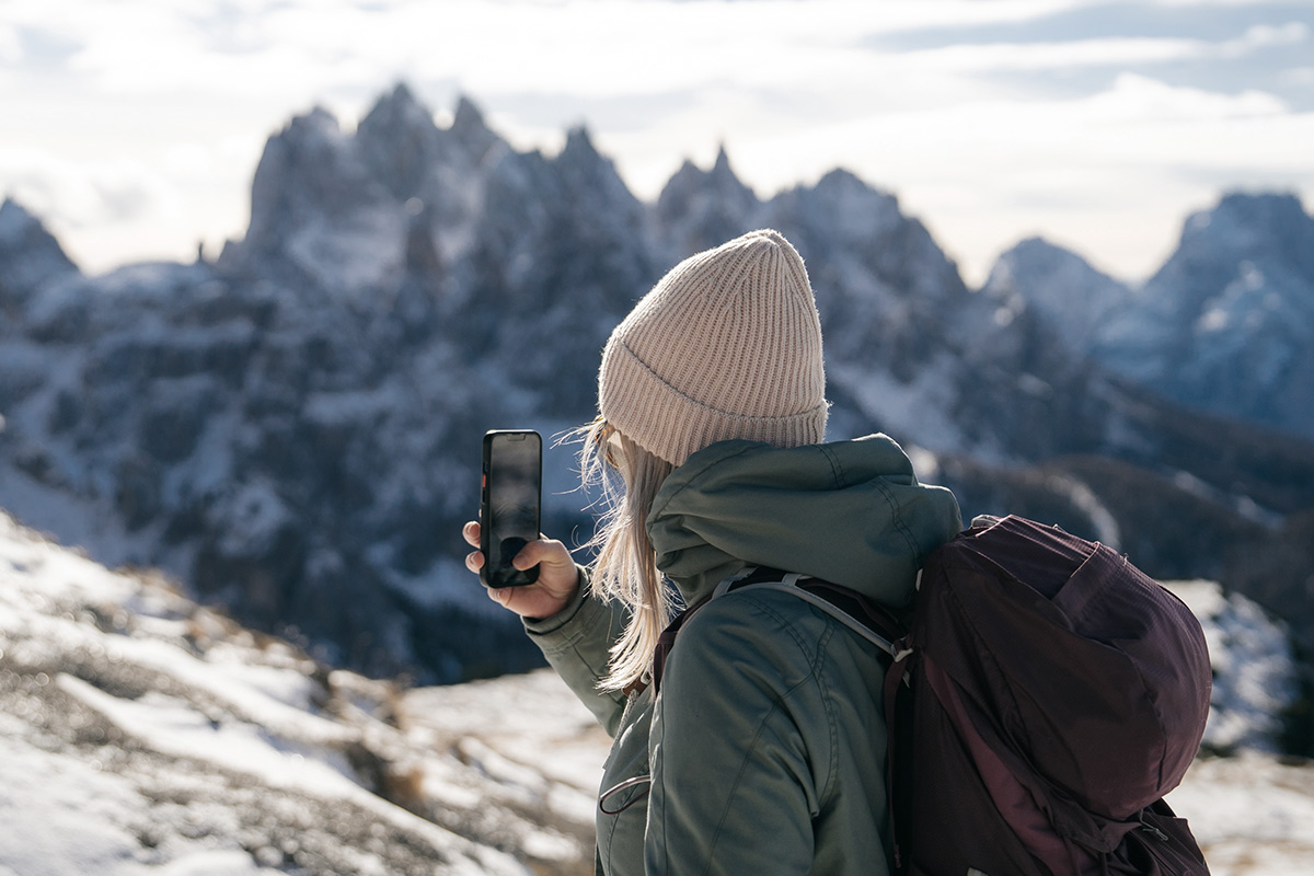 In Winterkleidung und mit Rucksack hält eine Frau in Hinterstoder die Berglandschaft mit ihrem Smartphone fest.