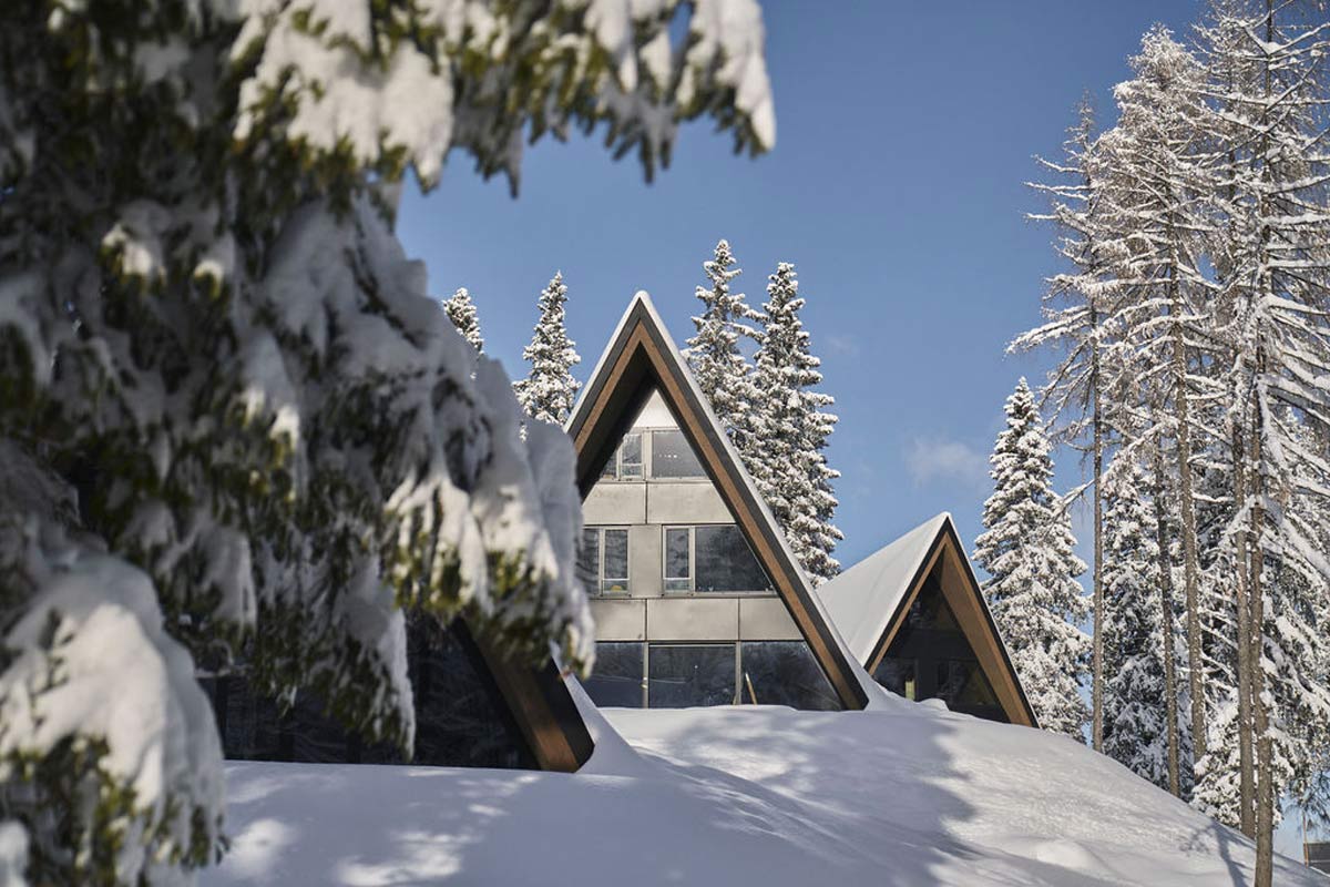Pures Bergidyll – unsere Chalets zwischen verschneiten Baumriesen. Dreieckiges Chalet des TRIFORÊT alpin.resort in Hinterstoder, umgeben von verschneiten Bäumen im Winter.