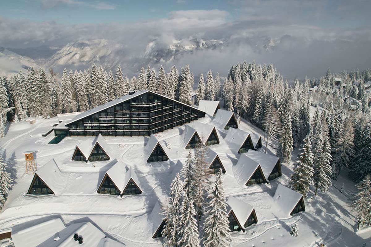 Winterwunder von oben – unser Resort im verschneiten Alpenpanorama. Luftaufnahme des verschneiten im TRIFORÊT alpin.resort mit Chalets und Hauptgebäude im Winterwald von Hinterstoder.
