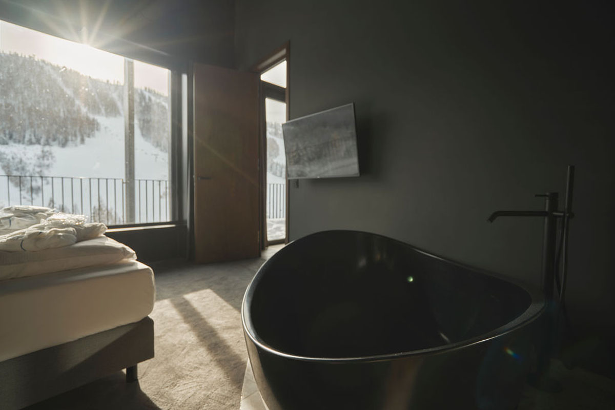 Entspannung pur mit Aussicht auf die Berge. Freistehende schwarze Badewanne im Zimmer mit Fensterblick auf verschneite Berghänge im TRIFORÊT alpin.resort.