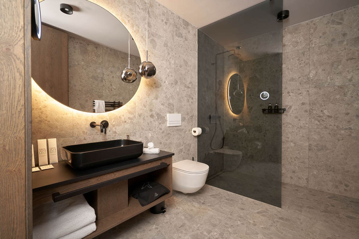 Für Ihre Morgenroutine mit Stil im Urlaub in den Alpen. Badezimmer mit Glasdusche, rundem Wandspiegel, Hänge-WC und dunklem Waschbecken im TRIFORÊT alpin.resort.