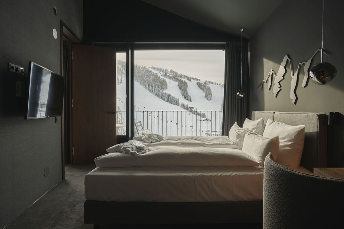 Einschlafen mit Aussicht in Ihrem Urlaub in den Alpen. Doppelbett Chalet des TRIFORÊT alpin.resort mit Wanddeko, Vorhängen und bodentiefem Fenster mit Bergblick.