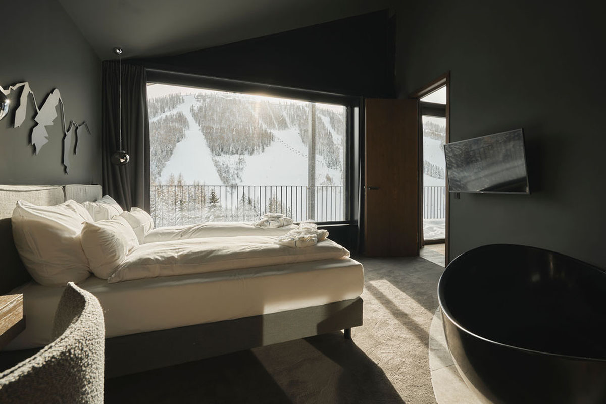 Aufwachen mit Panoramablick – Ihr Schlafzimmer im Urlaub in den Alpen. Moderner Schlafbereich mit großem Fenster und Blick auf verschneite Skipisten im TRIFORÊT alpin.resort in Hinterstoder.