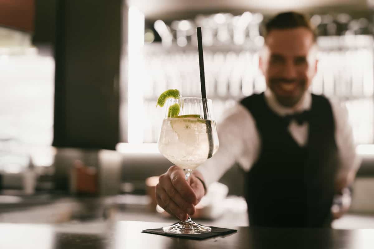 A bartender at TRIFORÊT alpin.resort hands an aperitif glass with lime over the bar counter for relaxation on vacation in the Alps.