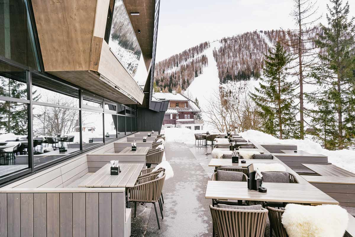 Wood-clad outdoor terrace of the Restaurant Hutterer surrounded by snow-covered trees and mountains in the TRIFORÊT alpin.resort.