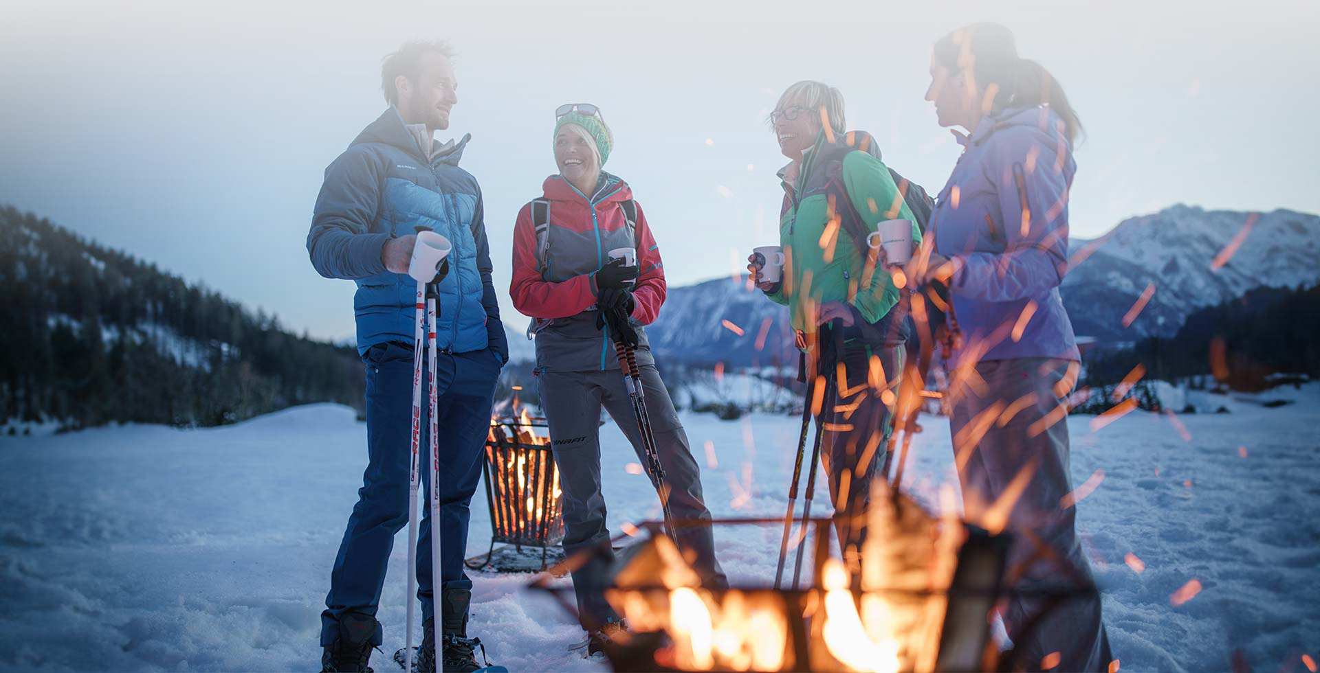 Group with a hot drink at the TRIFORÊT alpin.resorts snowshoeing event, standing by the fire in an alpine snowy landscape.