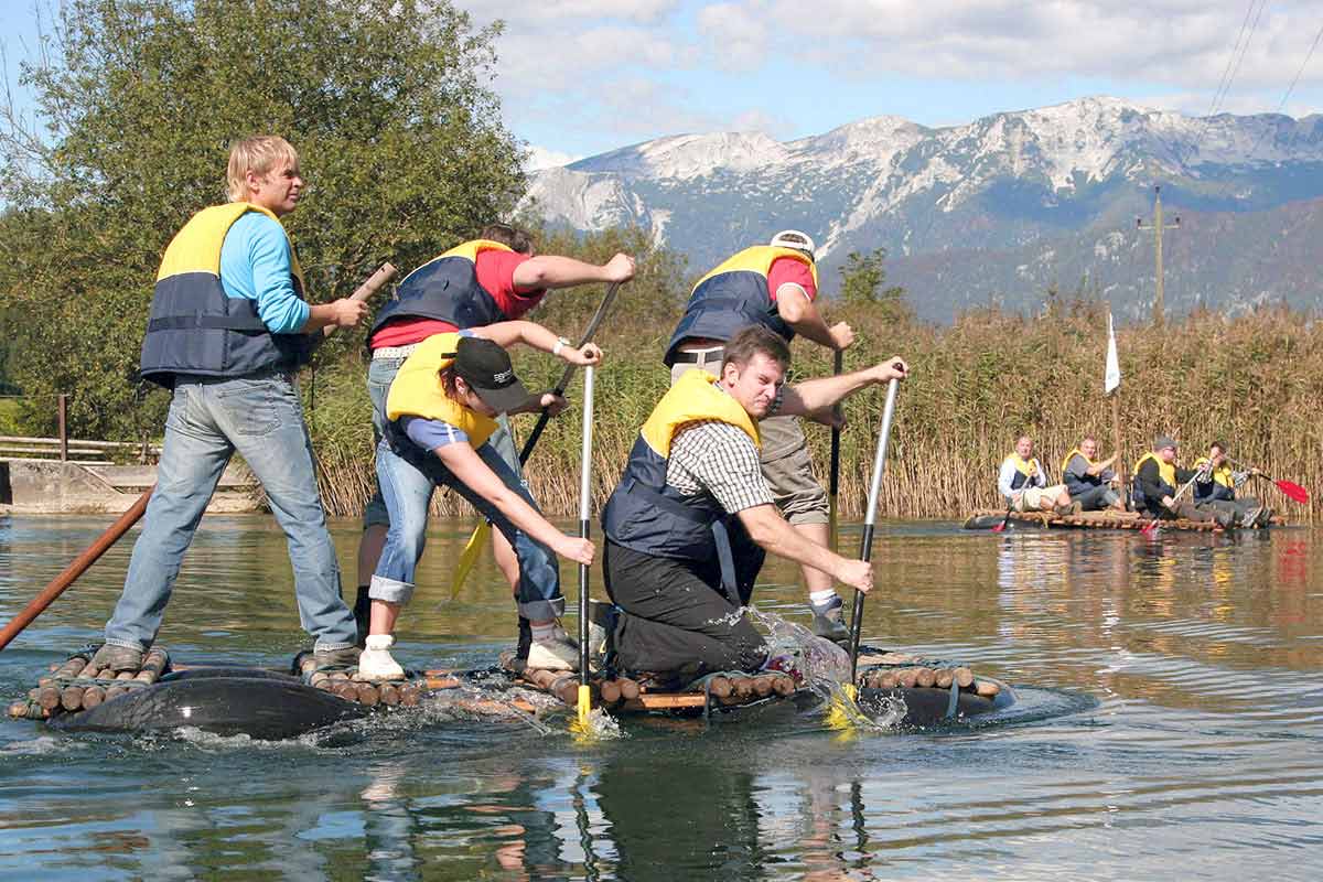 Several people paddle on a self-made raft in calm waters during a team building session at the TRIFORÊT alpin.resort.